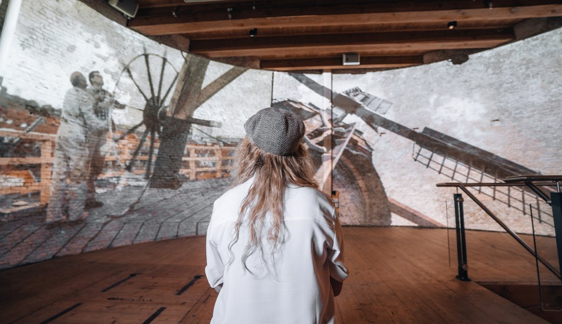 A person in a white shirt and beret looks at a historical photo projection of two men on top of a windmill. A person in a white shirt and beret looks at a historical photo projection of two men on top of a windmill.
