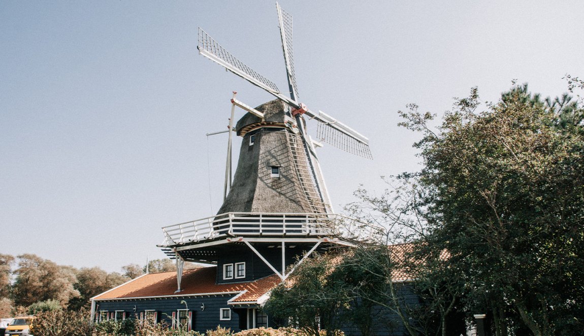 A classic Dutch windmill with a thatched roof and white sails, set against a clear sky, surrounded by greenery. A classic Dutch windmill with a thatched roof and white sails, set against a clear sky, surrounded by greenery.